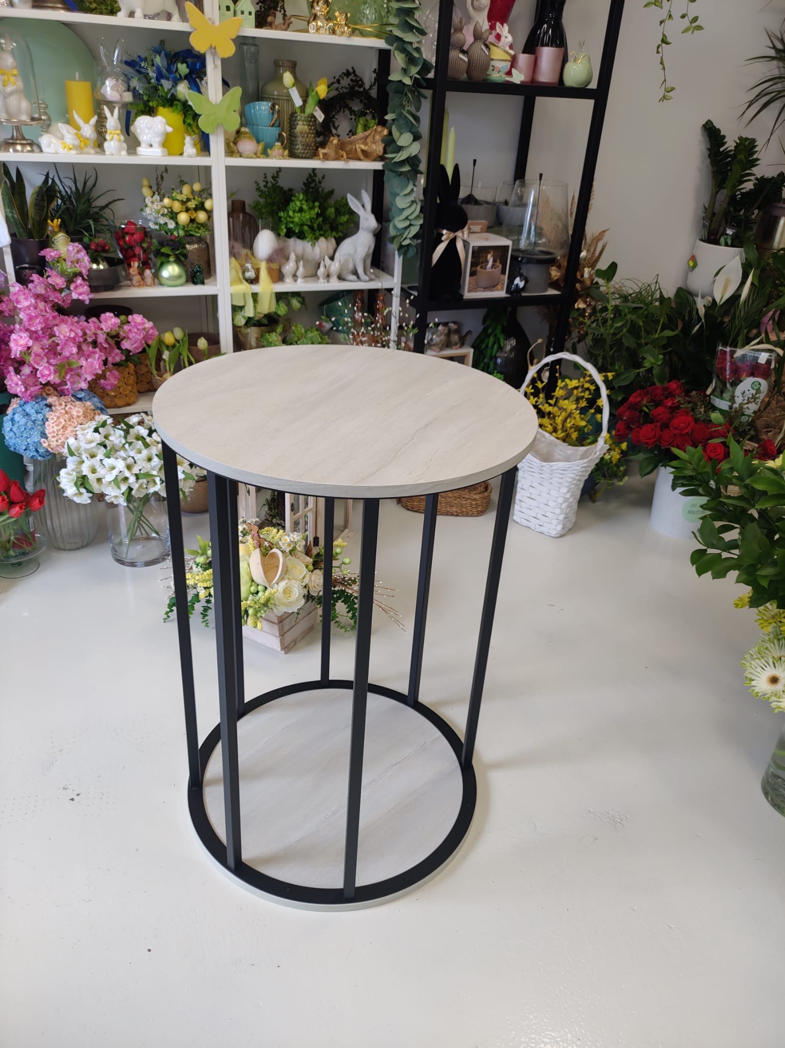 Okrągły stolik z jasnym blatem i czarną metalową podstawą w otoczeniu kwiatów i dekoracji, prezentowany w jasnym wnętrzu, prawdopodobnie kwiaciarni.