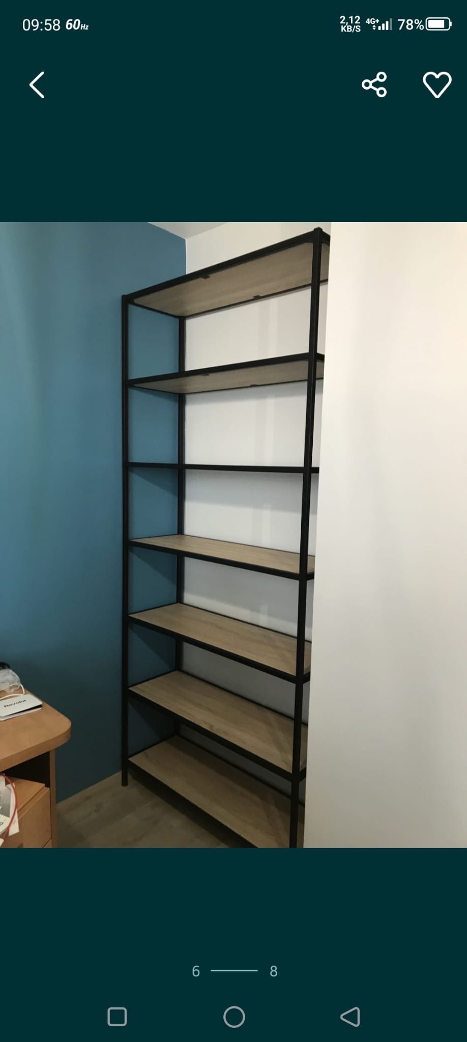 Metalowy regał z drewnianymi półkami na tle niebieskiej i białej ściany. Prosta konstrukcja, widoczne połączenia elementów. Minimalistyczny design.