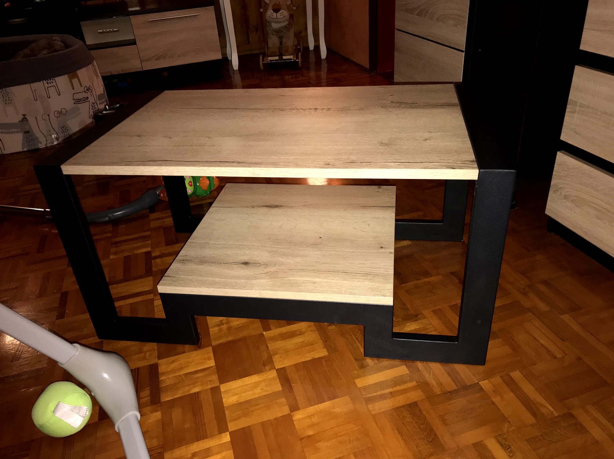 Stolik kawowy z blatem drewnopodobnym i czarnymi, metalowymi nogami, na tle parkietu. Widoczna dolna półka i fragmenty otoczenia.