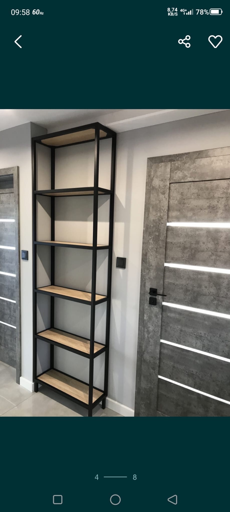 Wysoki regał z drewnianymi półkami i czarną, metalową ramą, stojący obok nowoczesnych drzwi z szarego betonu z podłużnymi, białymi paskami.