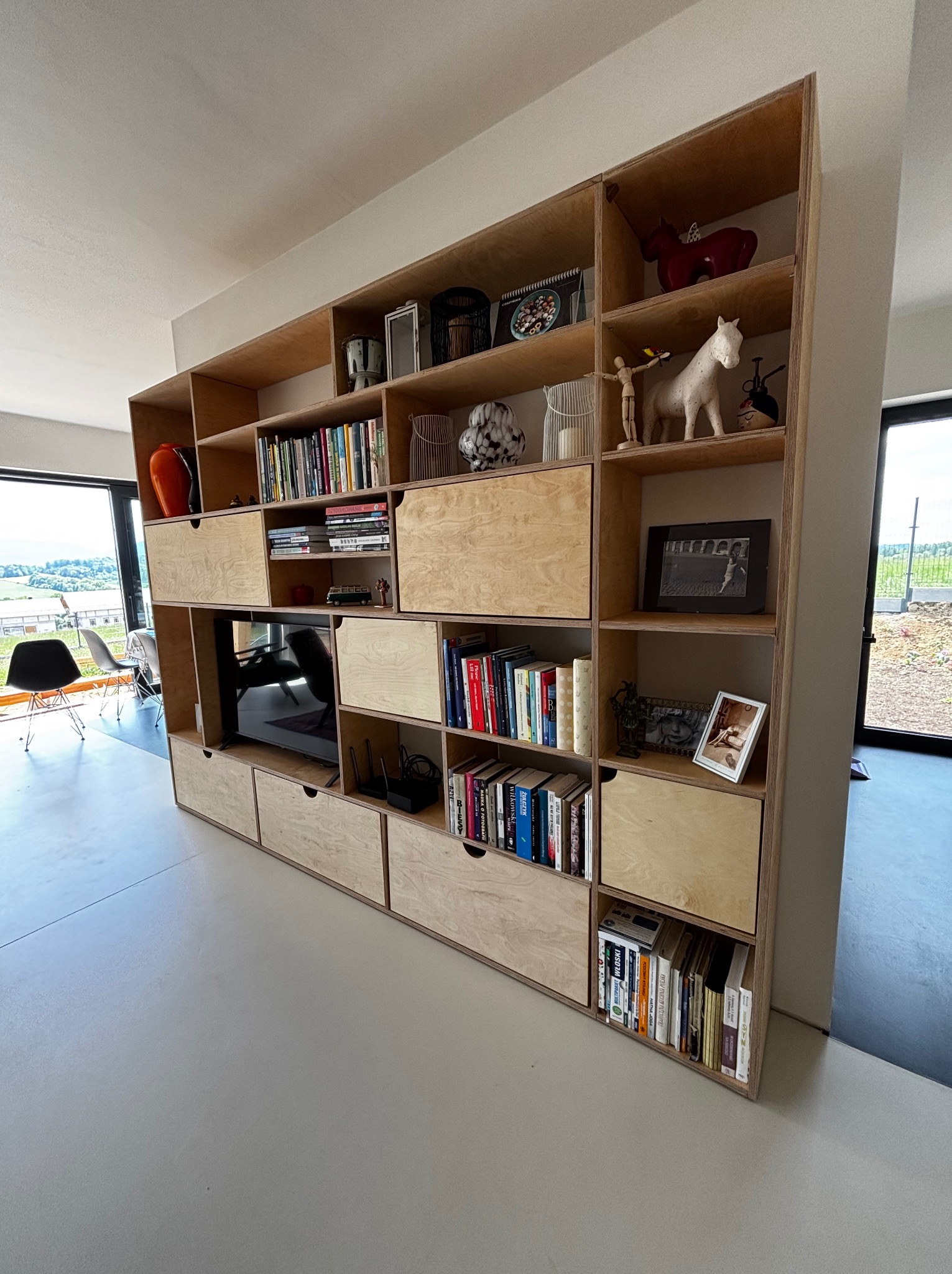 Półka z jasnej sklejki, wbudowana w ścianę, z licznymi przegródkami, książkami i dekoracjami. W środkowej części wnęka na telewizor. Nowoczesny minimalizm.