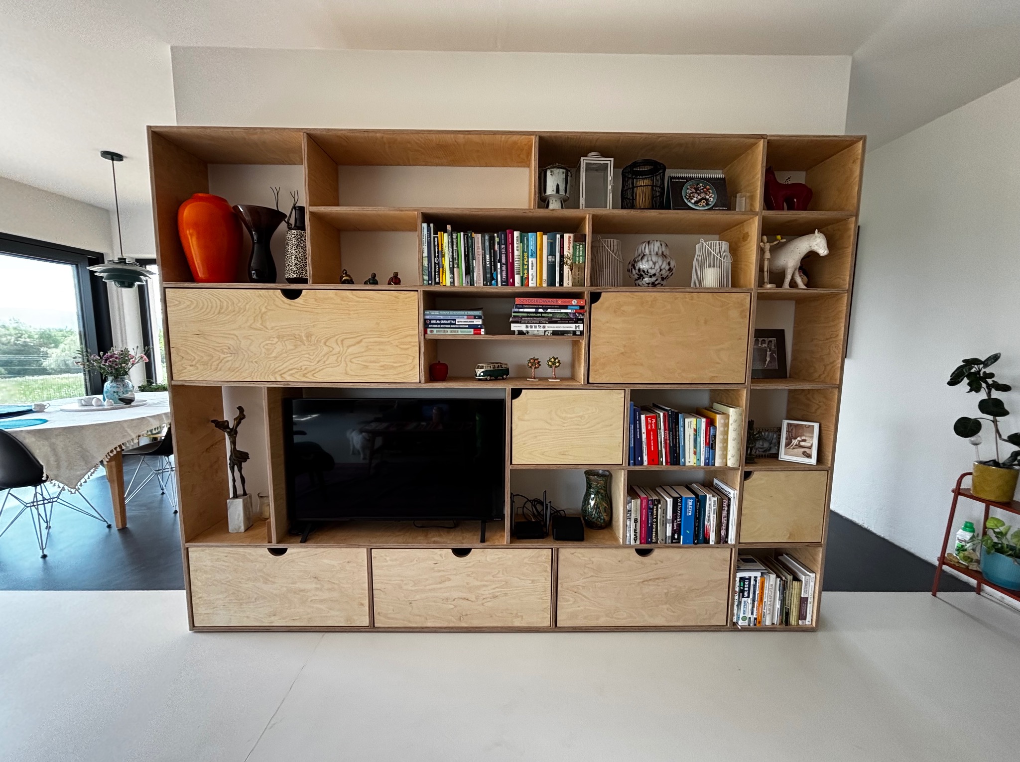 Duża, drewniana szafa z półkami i szufladami z jasnej sklejki, w której znajduje się telewizor, książki i dekoracje. Szafa stoi na tle białej ściany.