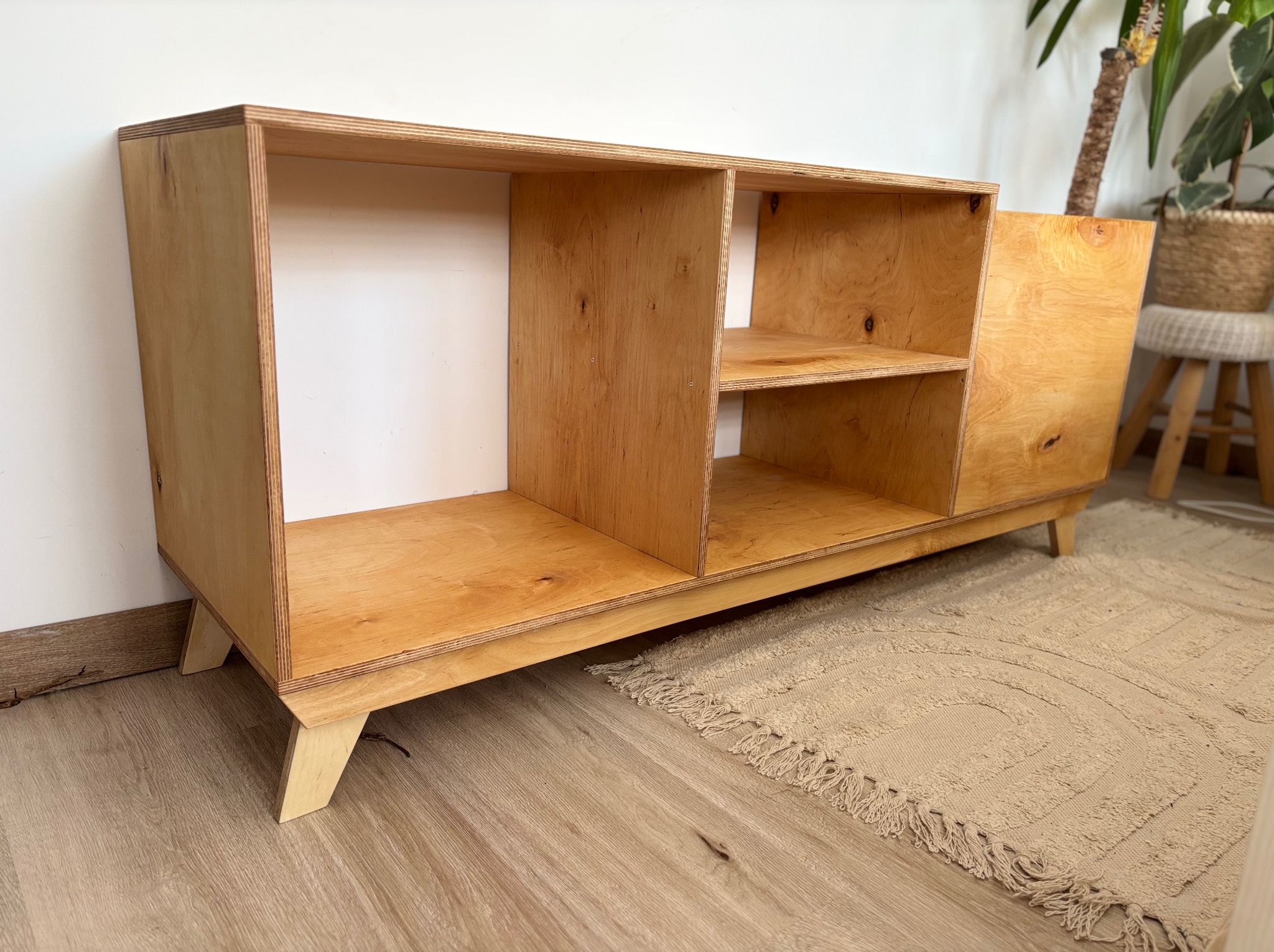 Drewniana komoda z półkami z jasnej sklejki, na niskich nóżkach, w minimalistycznym stylu. W tle widoczny dywan i roślina doniczkowa.