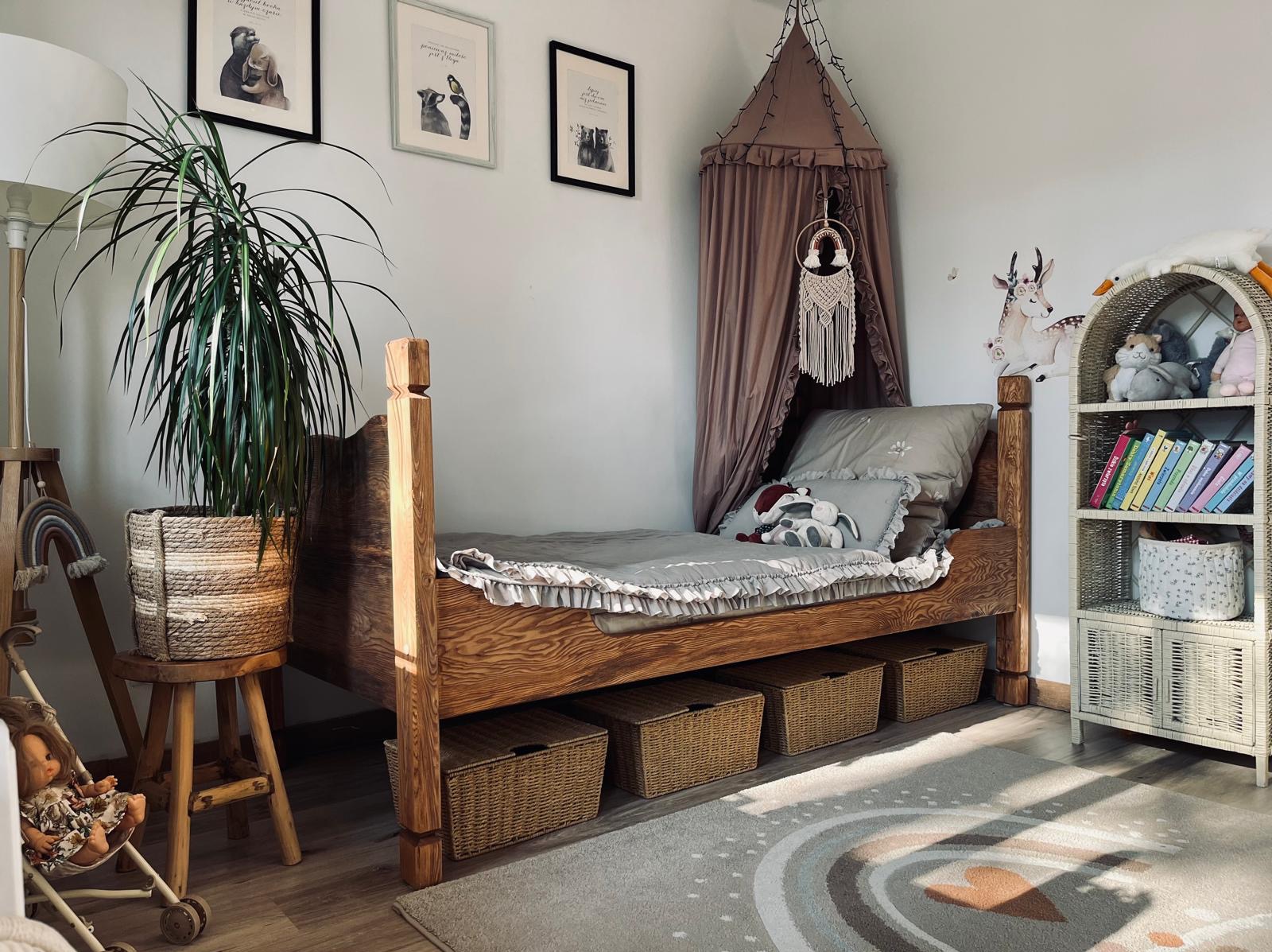Drewniane łóżko dziecięce z baldachimem w pokoju, z trzema koszami pod spodem. Na ścianach wiszą obrazki i naklejki. Obok stoi regał z książkami.