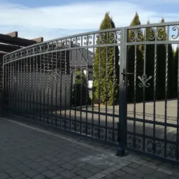 Metalowa brama wjazdowa z ozdobnymi elementami, widoczna z zewnątrz posesji, z tłem zielonych drzew i domu z ciemnym dachem.