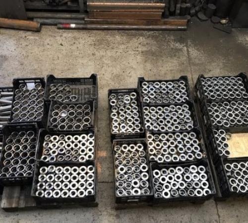 Seryjne metalowe tuleje w czarnych plastikowych skrzynkach, widoczne ślady obróbki, w tle metalowe rury