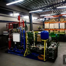 Kompleksowa instalacja przemysłowa z rurami w kolorach czerwonym, niebieskim i zielonym, zbiornikiem, oraz elementami sterującymi, umieszczona w hali produkcyjnej z regałami magazynowymi w tle.