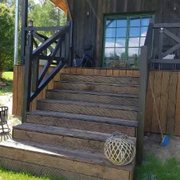 Drewniane schody prowadzące na taras domu z ciemnego drewna, widoczne dekoracyjne lampiony i elementy wykończenia w czarnym kolorze.