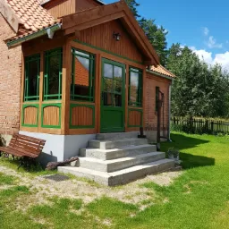 Ceglany dom z drewnianą werandą pomalowaną na zielono, betonowe schody wejściowe, drewniana ławka i zadbany trawnik w słoneczny dzień.