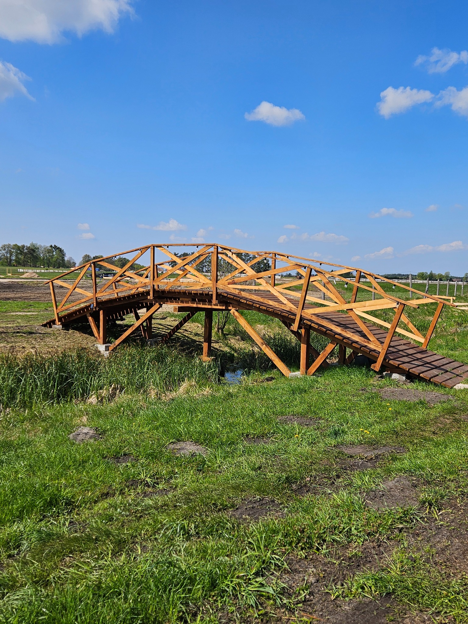 Drewniany mostek ogrodowy z ażurową konstrukcją, prowadzący nad niewielkim ciekiem wodnym, w tle zielona trawa i błękitne niebo.