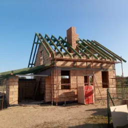 Surowy stan budowy domu z cegły, widoczna drewniana konstrukcja dachu, komin z cegły, rusztowania i palety z cegłami Wienerberger w słoneczny dzień.