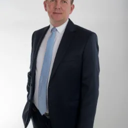 Portret mężczyzny w granatowym garniturze i błękitnym krawacie na jasnoszarym tle, z ręką w kieszeni.