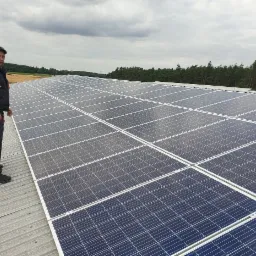 Instalator w kombinezonie roboczym stoi na dachu z blachy trapezowej, obok rzędu paneli słonecznych w słoneczny dzień. W tle widać pola i las.