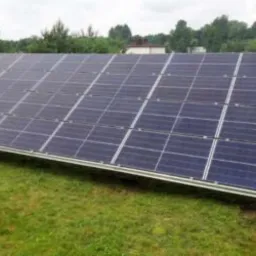 Panele fotowoltaiczne zamontowane na trawniku, widoczne elementy konstrukcyjne, w tle drzewa i dach domu.