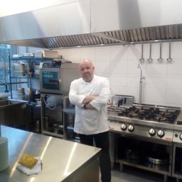CHEF UNITED - Szef kuchni w białym uniformie stoi na tle profesjonalnej, stalowej kuchni z okapem, kuchenką gazową, piekarnikiem konwekcyjnym i akcesoriami kuchennymi.