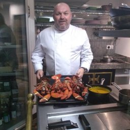 CHEF UNITED - Szef kuchni w białym uniformie prezentuje tacę z czterema ugotowanymi homarami obok garnka z żółtym sosem na płycie kuchennej w profesjonalnej kuchni.