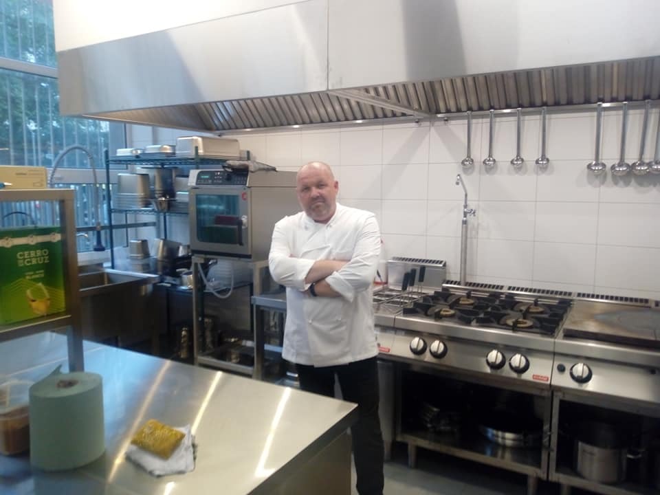 Szef kuchni w białym uniformie stoi na tle profesjonalnej, stalowej kuchni z okapem, kuchenką gazową, piekarnikiem konwekcyjnym i akcesoriami kuchennymi.