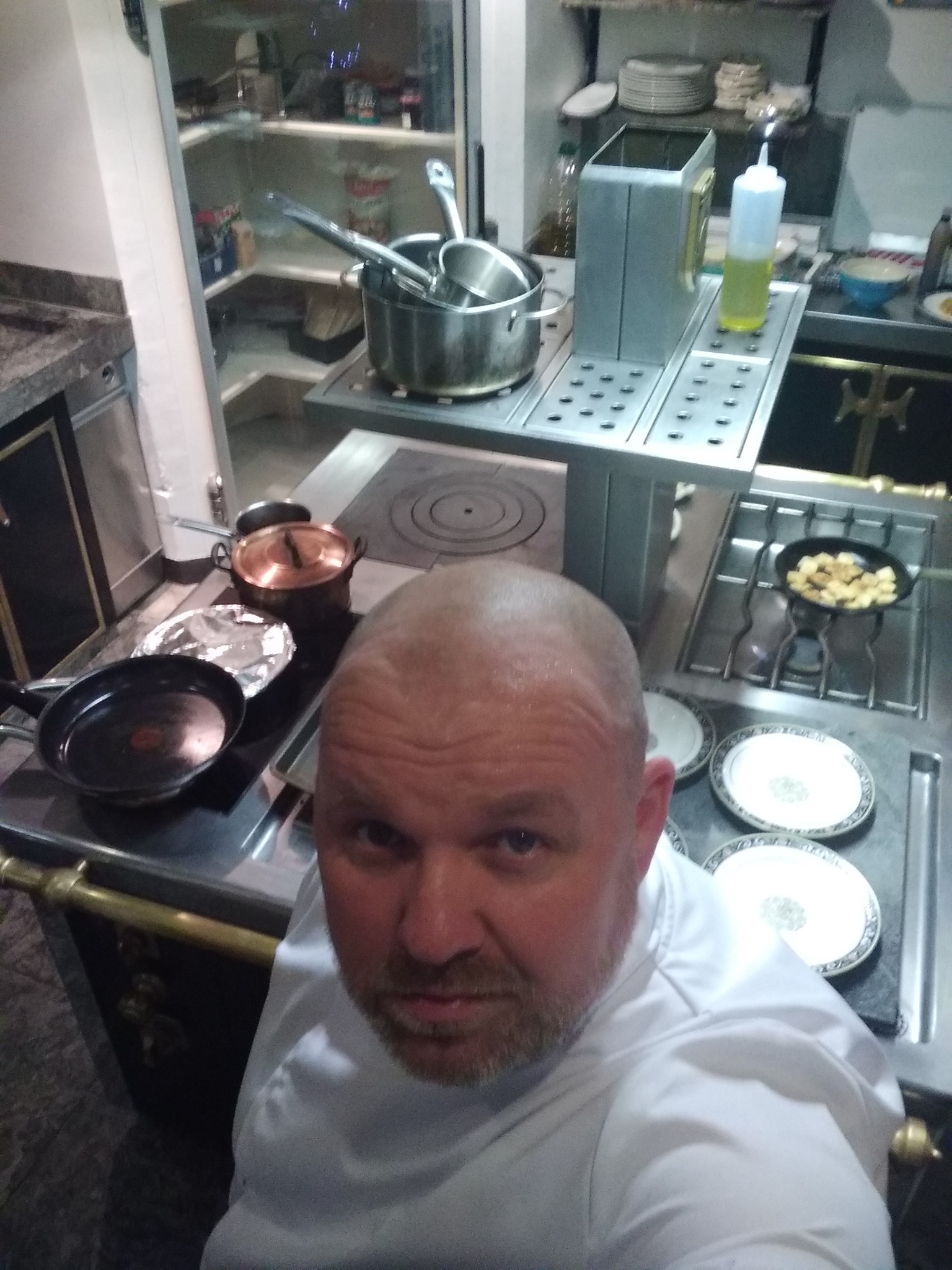 Selfie kucharza w białym uniformie na tle kuchni z profesjonalnym sprzętem: garnkami, patelniami i talerzami, z widoczną kuchenką gazową i półkami z produktami.