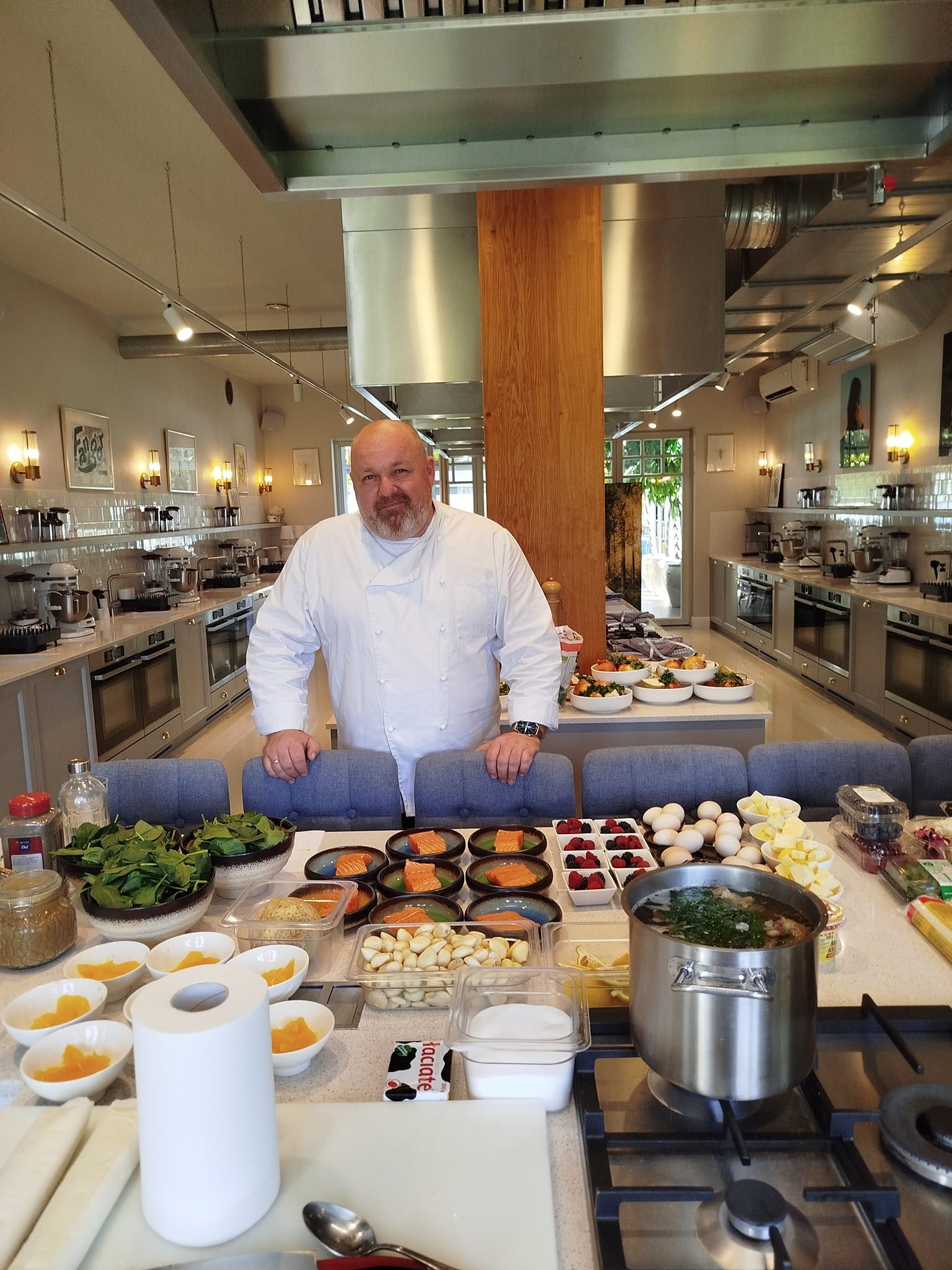 Kucharz w białym uniformie prezentuje stół z przygotowanymi składnikami: łosoś, owoce, warzywa i bulion, w tle widoczna profesjonalna kuchnia z licznymi stanowiskami pracy.