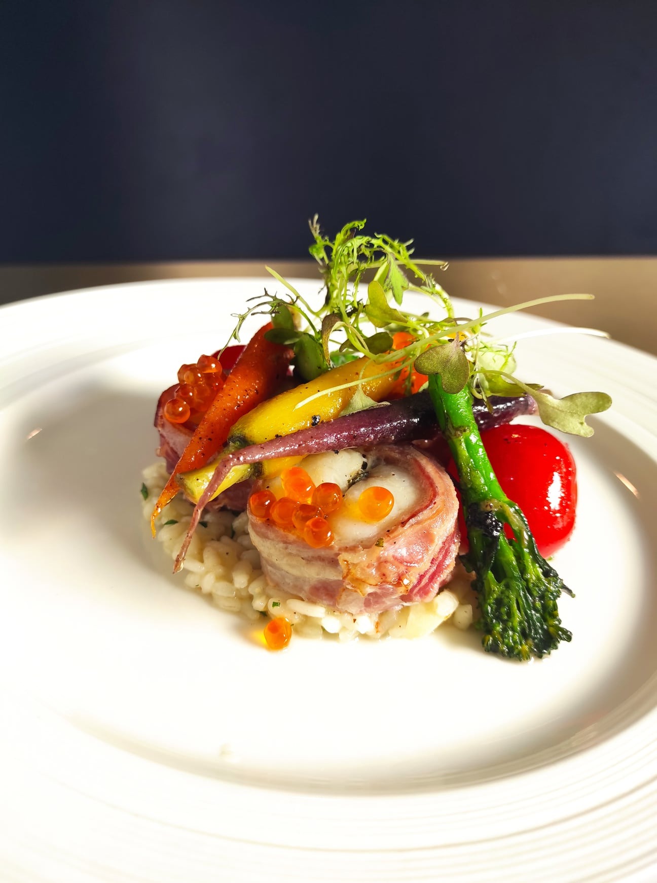 Elegancko podane danie: ryba zawinięta w bekon na risotto, udekorowana kolorowymi warzywami, kawiorem i zieleniną na białym talerzu.