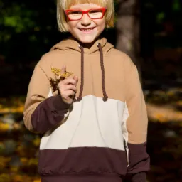Uśmiechnięty chłopiec w bluzie z kapturem w kolorach ziemi i czerwonych okularach, trzymający w palcach uschnięty liść na tle jesiennego parku.