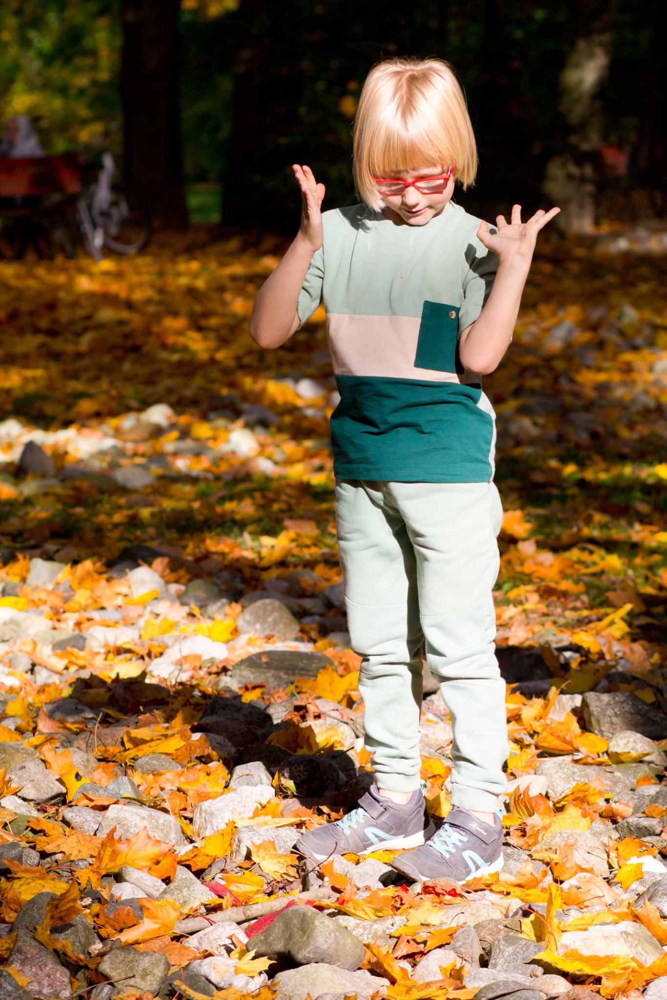 Blondwłosy chłopiec w okularach i nowoczesnym ubraniu stoi na kamieniach pokrytych jesiennymi liśćmi, prezentując strój z bluzką w bloki kolorów i spodniami dresowymi.