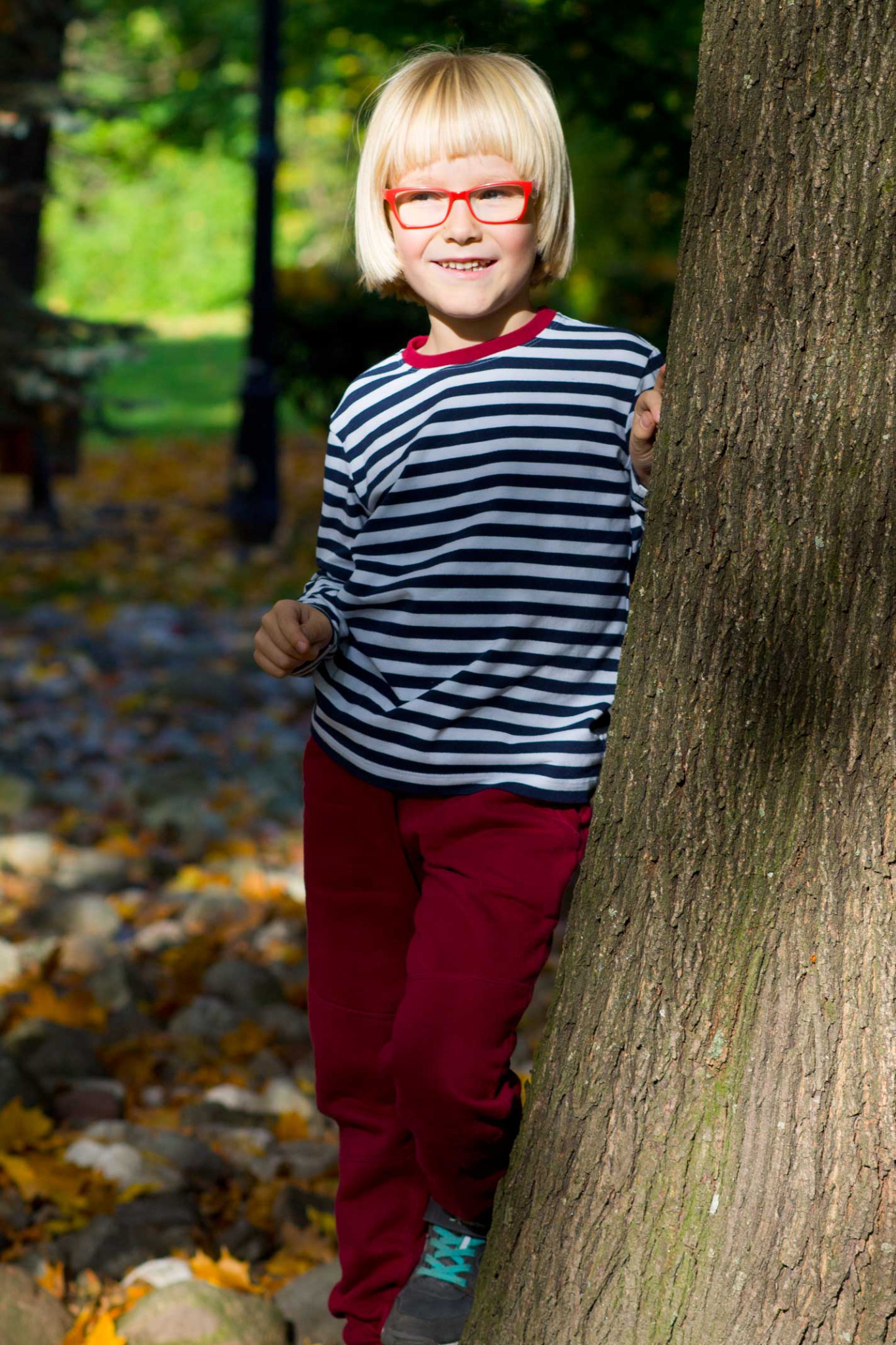 Uśmiechnięty chłopiec z blond grzywką i w czerwonych okularach, ubrany w pasiasy sweter i czerwone spodnie, opiera się o drzewo w jesiennym parku.