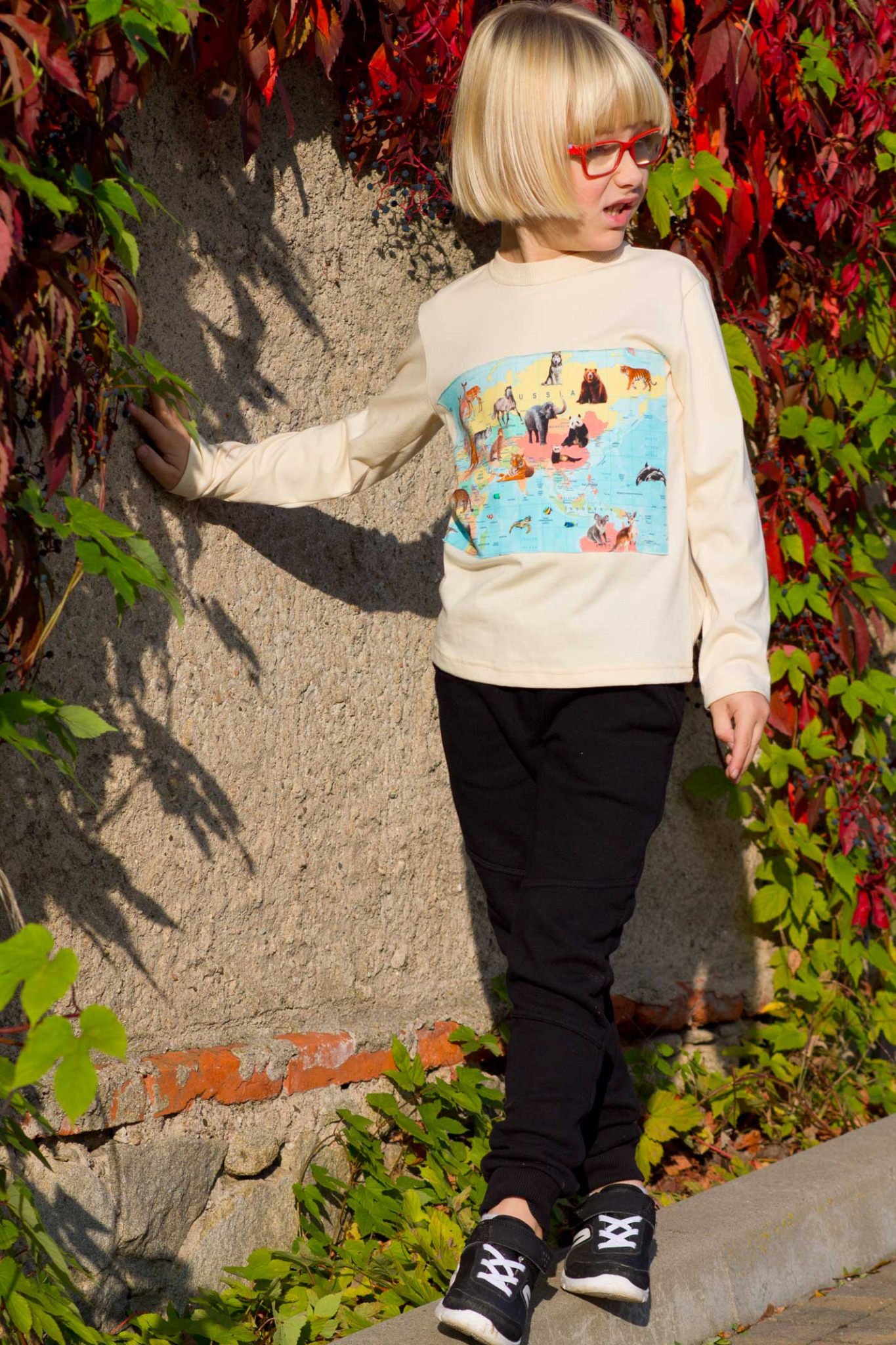 Chłopiec w okularach i koszulce z mapą świata i zwierzętami, pozuje na tle ściany porośniętej bluszczem.