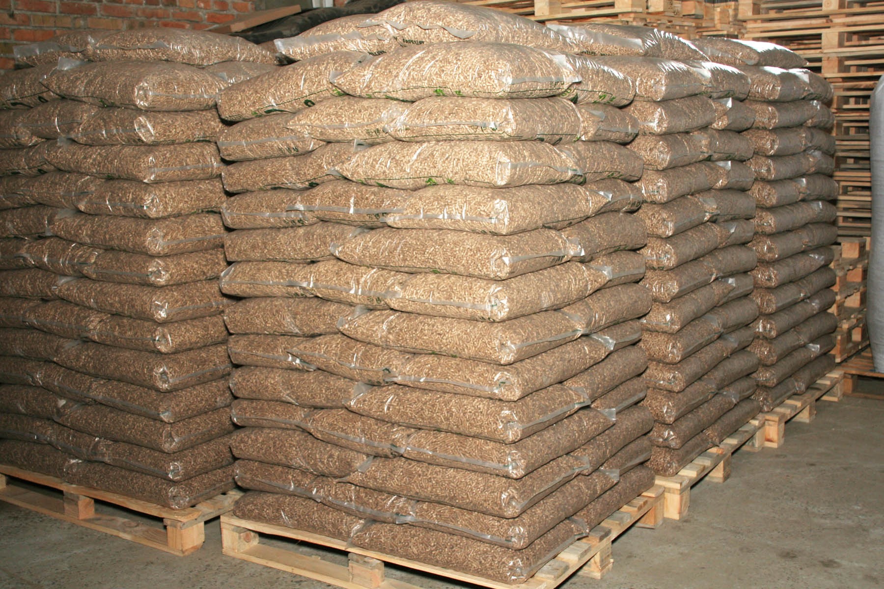 Stosy worków z pelletem ułożone na drewnianych paletach w magazynie z ceglaną ścianą w tle.