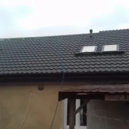 Dom z częściowo wykończonym dachem pokrytym ciemną dachówką, widoczne okna dachowe i fragment konstrukcji dachu przed ułożeniem pełnego pokrycia.