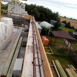 Budowa domu jednorodzinnego: widok z góry na zbrojenie fundamentów w szalunku, w tle pustaki, drewniane stemple i elementy konstrukcji więźby dachowej oraz budynki gospodarcze i zieleń.