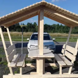 Drewniany stół piknikowy z ławkami pod zadaszeniem, surowe drewno, w tle biały samochód i krajobraz polny.