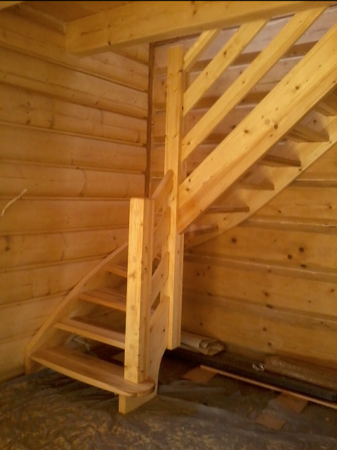 Drewniane schody z balustradą w surowym stanie wewnątrz drewnianego domu z bali, widoczne niedoskonałości obróbki i montażu.