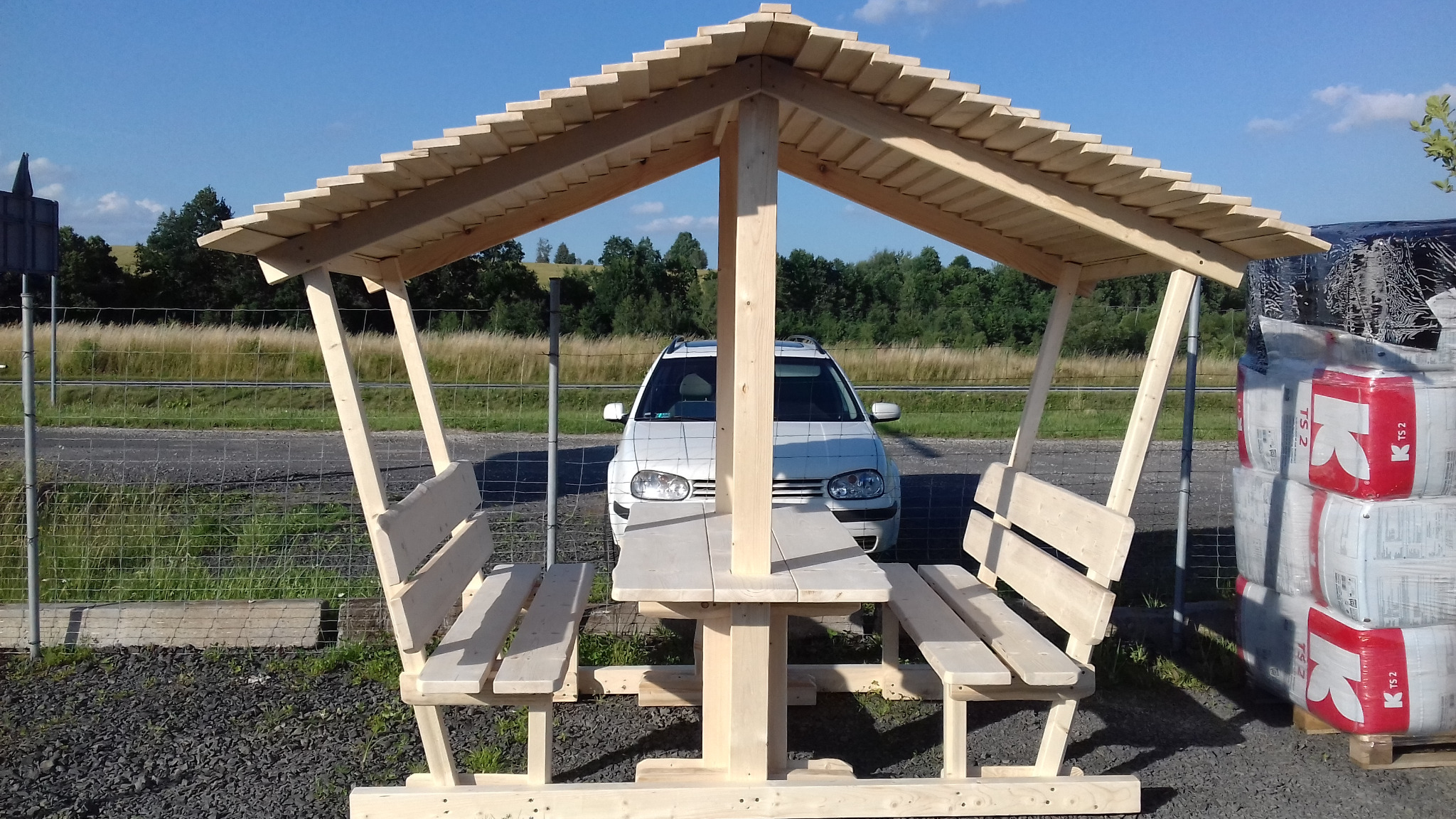 Drewniany stół piknikowy z ławkami pod zadaszeniem, surowe drewno, w tle biały samochód i krajobraz polny.