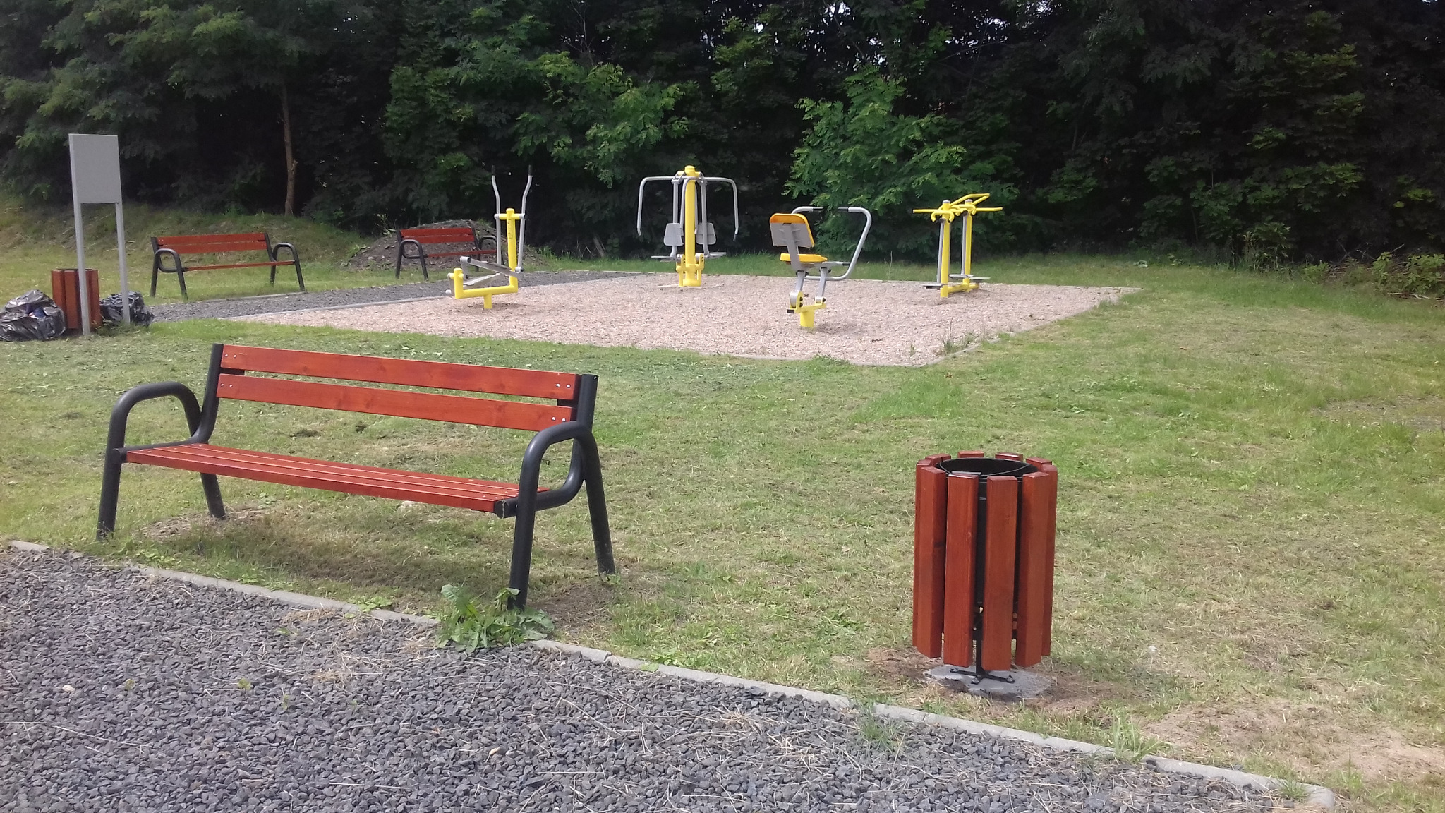 Plenerowa siłownia w Mirsku z drewnianymi ławkami i koszami na śmieci, otoczona zielenią.
