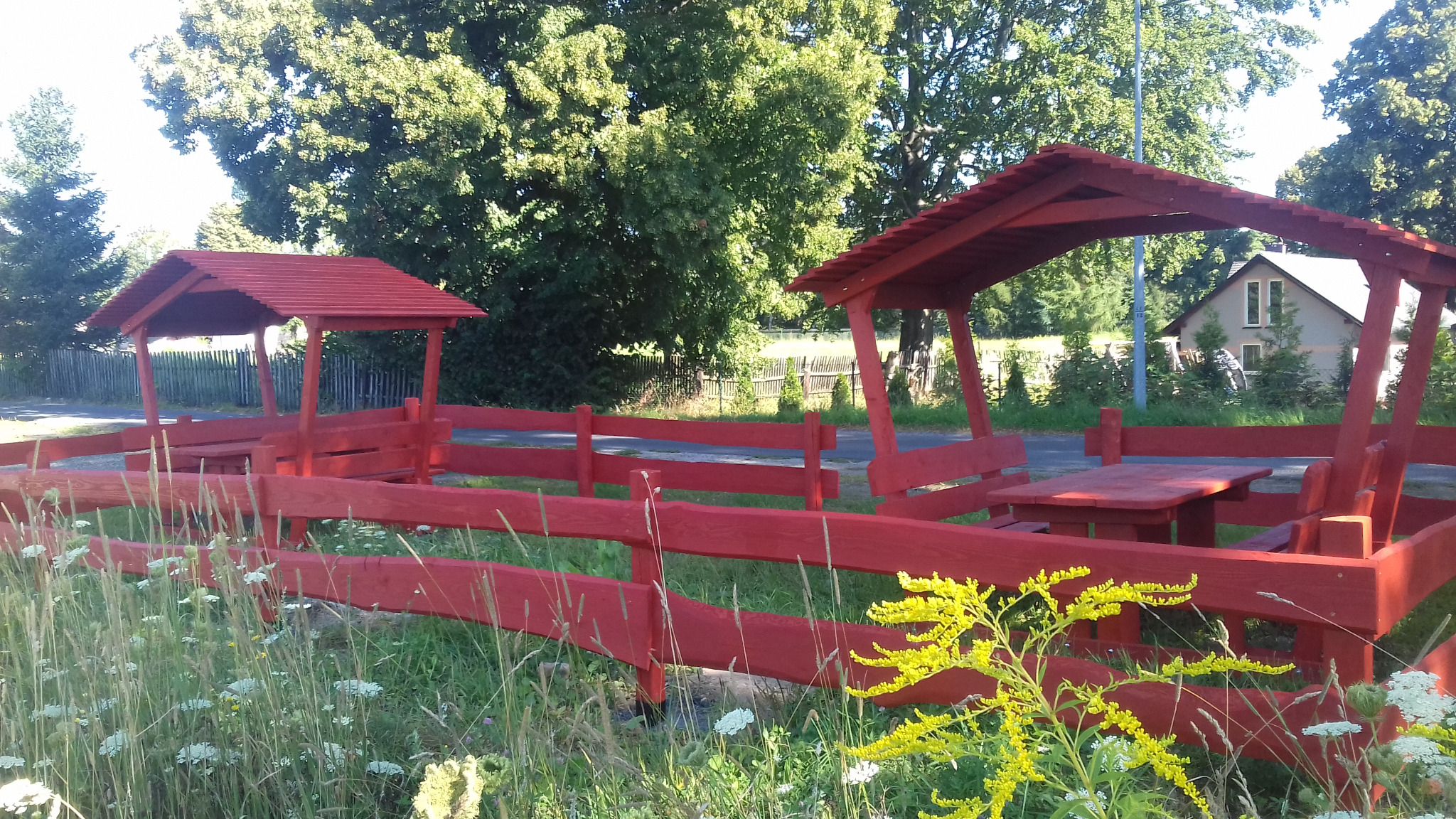 Czerwona drewniana altana z ławkami i stołem w plenerze, otoczona ogrodzeniem i zielenią, widok na dom w tle.