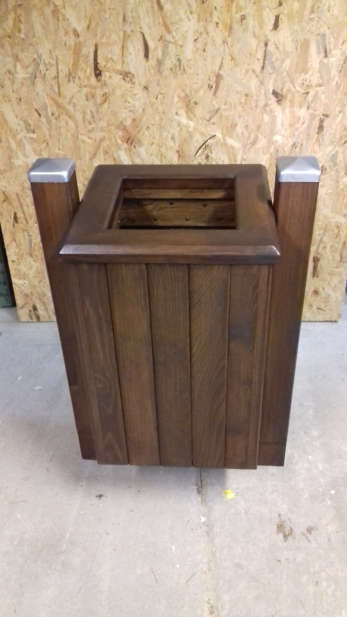 Kwadratowa, drewniana donica z ciemnego drewna z metalowymi nakładkami na rogach, widoczna na tle ściany z płyty OSB i betonowej podłogi.