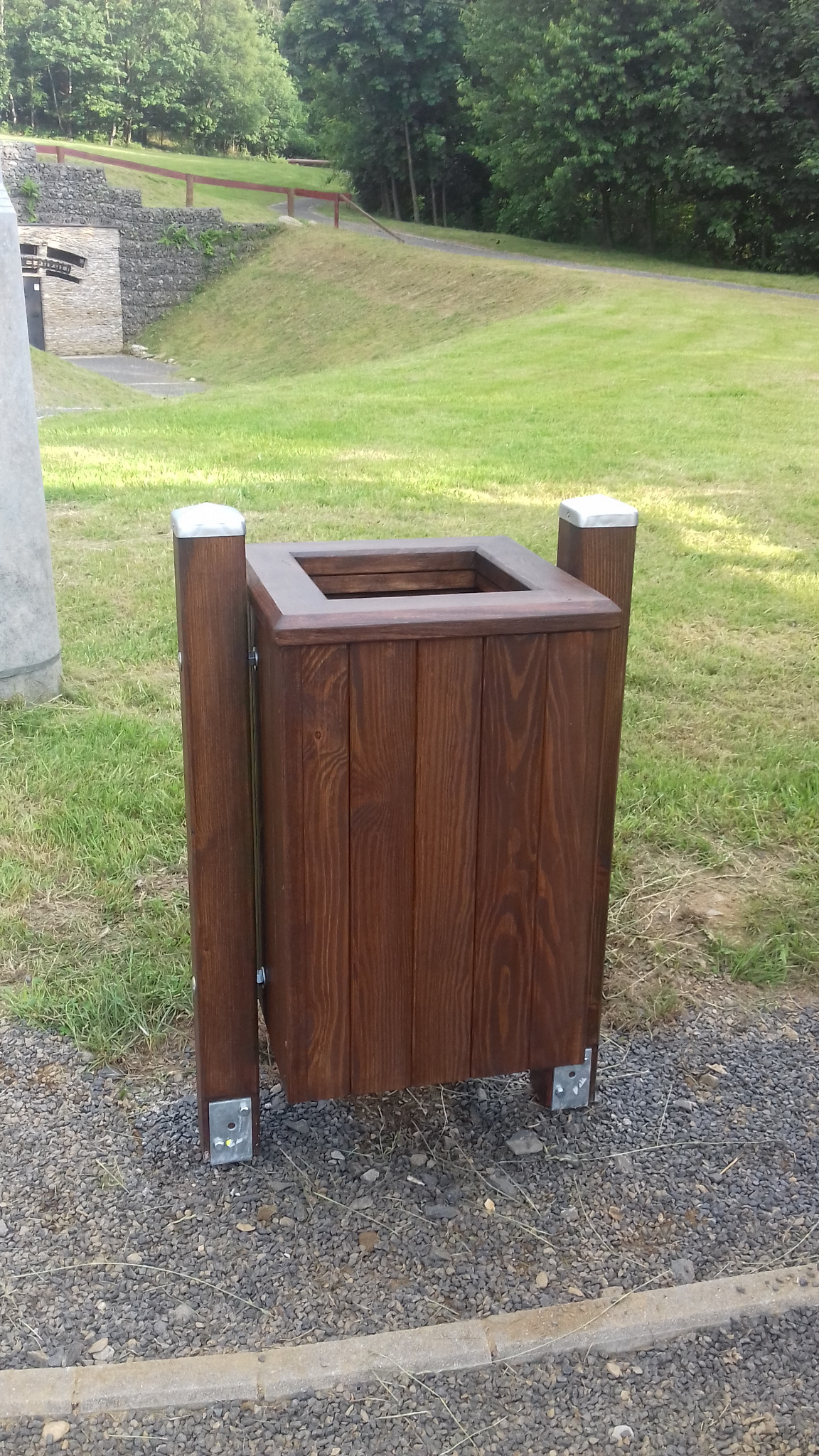 Brązowy, drewniany kosz na śmieci z metalowymi elementami, umieszczony na żwirowym podłożu w otoczeniu zieleni.