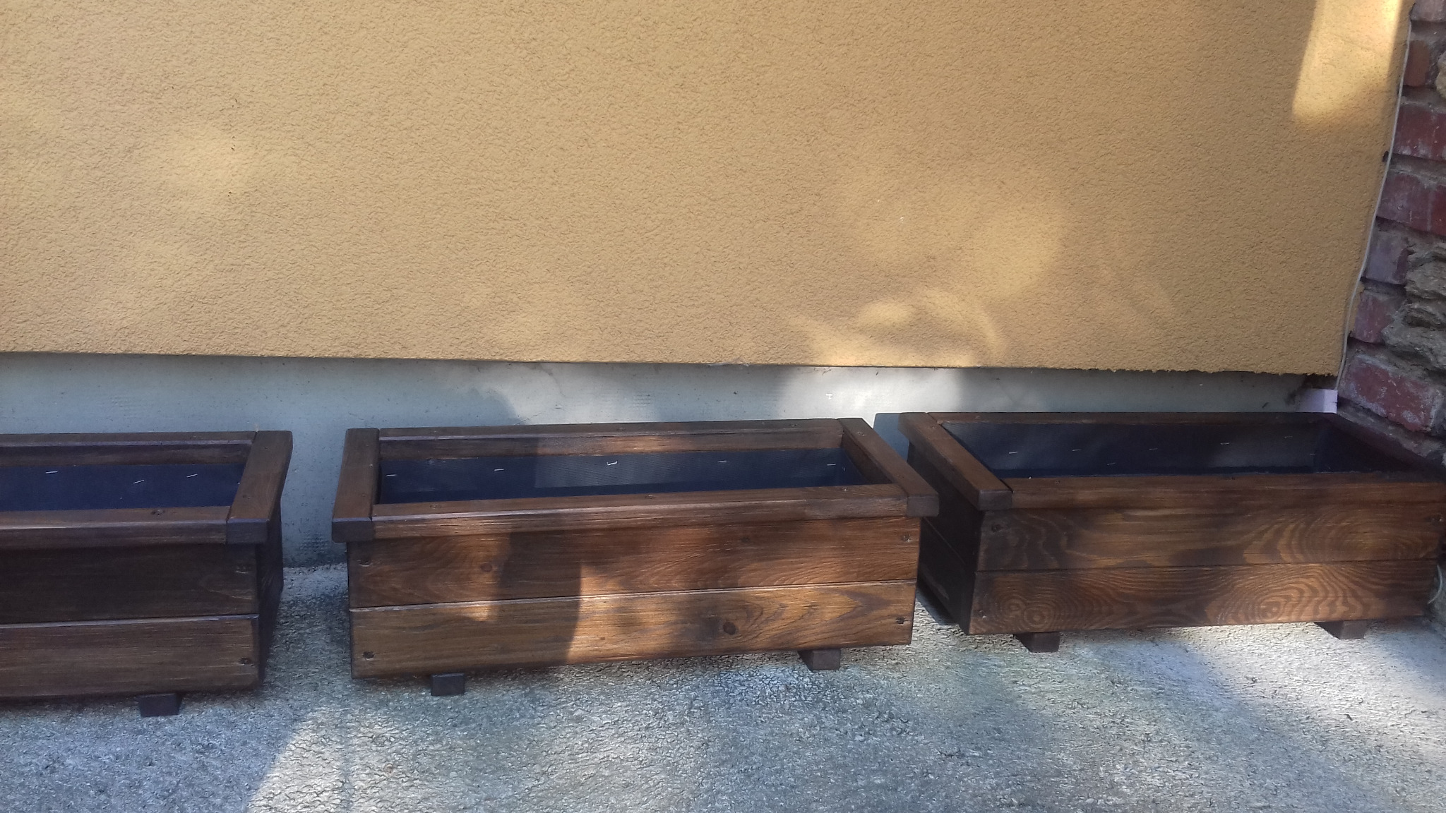 Trzy ciemnobrązowe, drewniane skrzynki na kwiaty/zioła stojące przy ścianie budynku z żółtym tynkiem i fragmentem muru z czerwonej cegły.