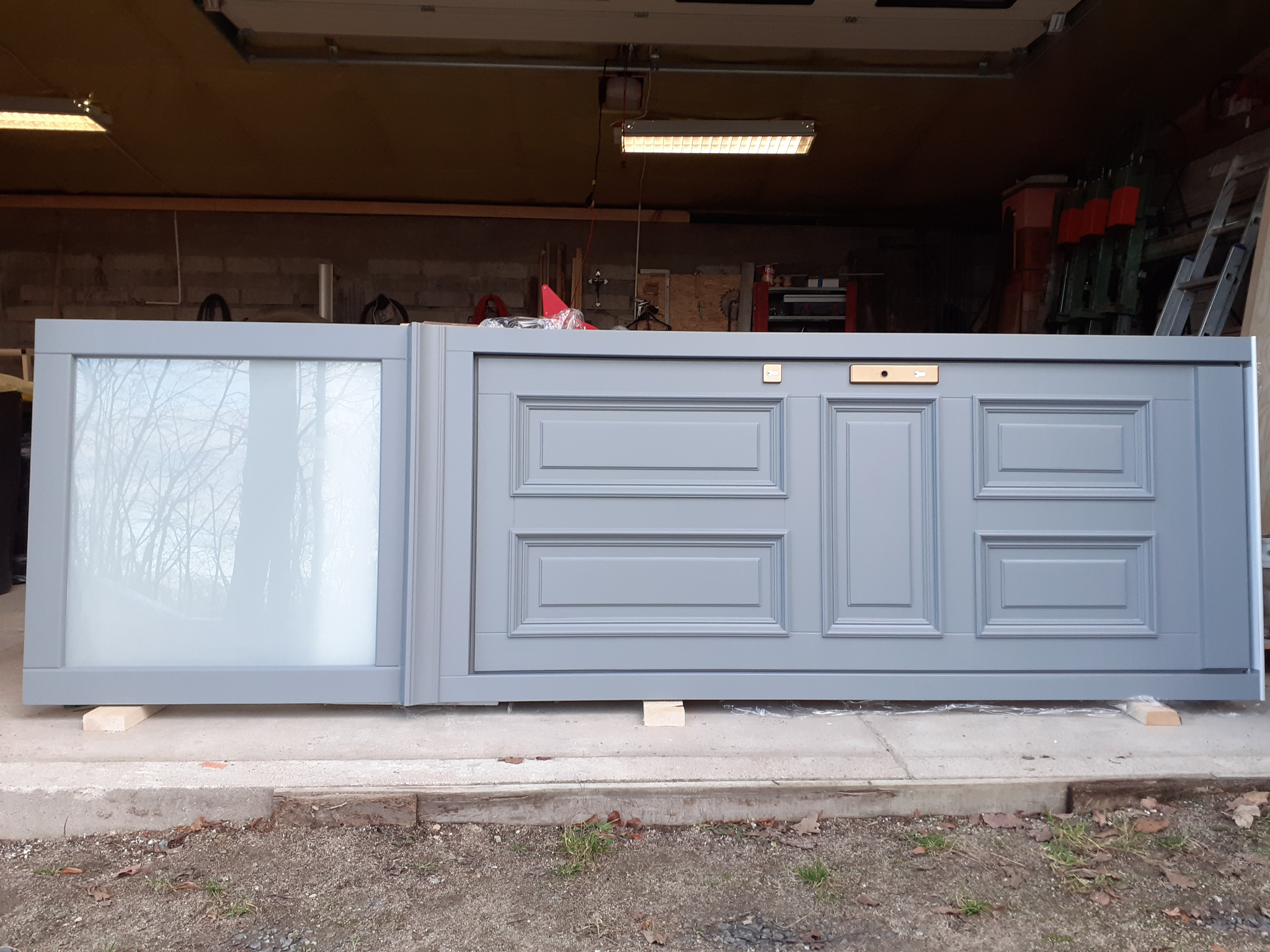 Szary, częściowo przeszklony front szafy z dekoracyjnymi panelami, oparty na drewnianych klockach w garażu.