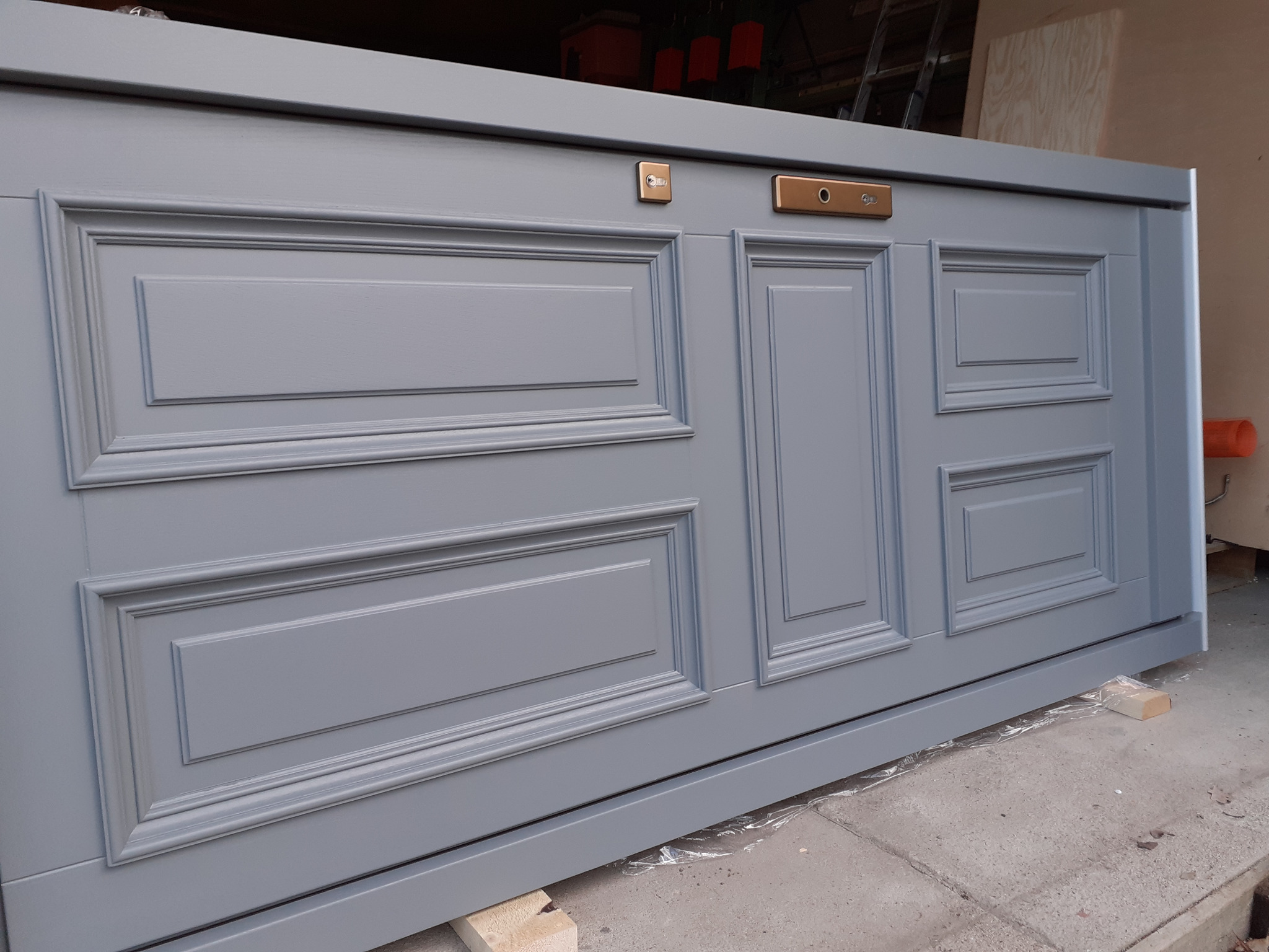 Szare, lakierowane drzwi z panelami dekoracyjnymi i mosiężnymi okuciami, wsparte na drewnianych klockach podczas montażu.