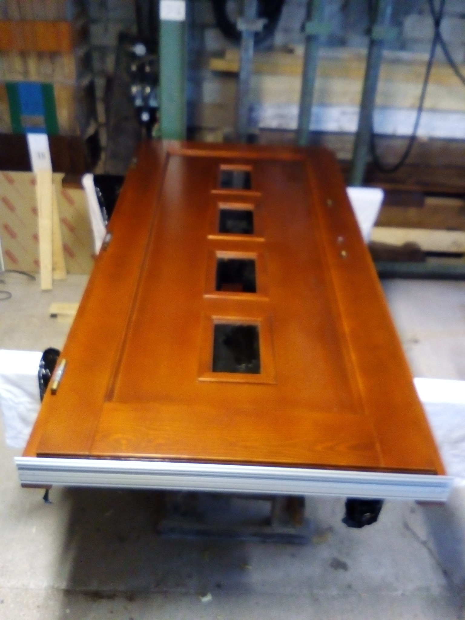 Lakierowane na brązowo drzwi z trzema kwadratowymi przeszkleniami, leżące na podkładkach w warsztacie.