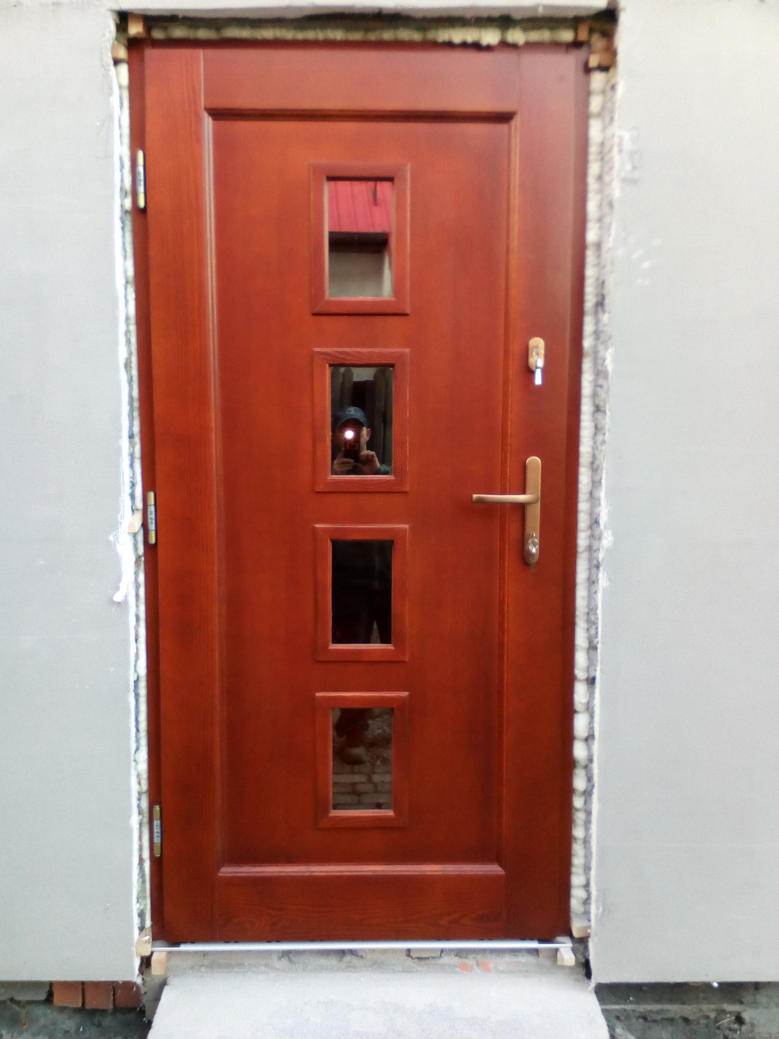 Ciemnobrązowe drzwi wejściowe z czterema małymi okienkami, widoczne ślady montażu, odbicie osoby robiącej zdjęcie w środkowym okienku.