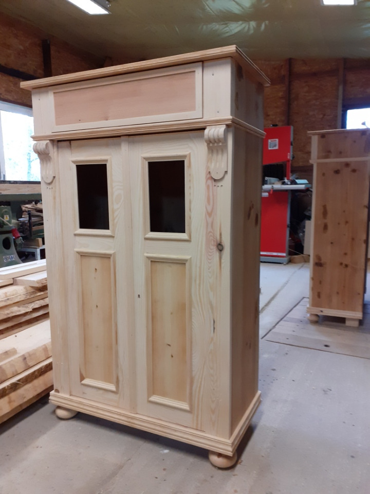 Niedokończona, drewniana szafa z dwoma drzwiami z prostokątnymi wgłębieniami, ozdobnymi elementami po bokach i kulistymi nóżkami, widoczna w warsztacie stolarskim.