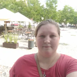 Portret kobiety w czerwonej koszulce z kwiatowym wzorem, na tle letniego ogródka kawiarnianego z białymi parasolami i zielonymi drzewami.