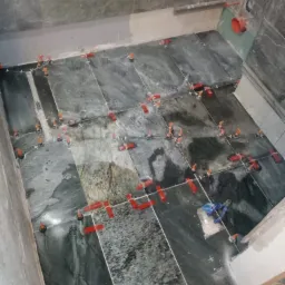 Układanie ciemnych, marmurkowych płytek podłogowych w łazience, z systemem poziomowania płytek (czerwone kliny i pomarańczowe dystanse) w trakcie prac.