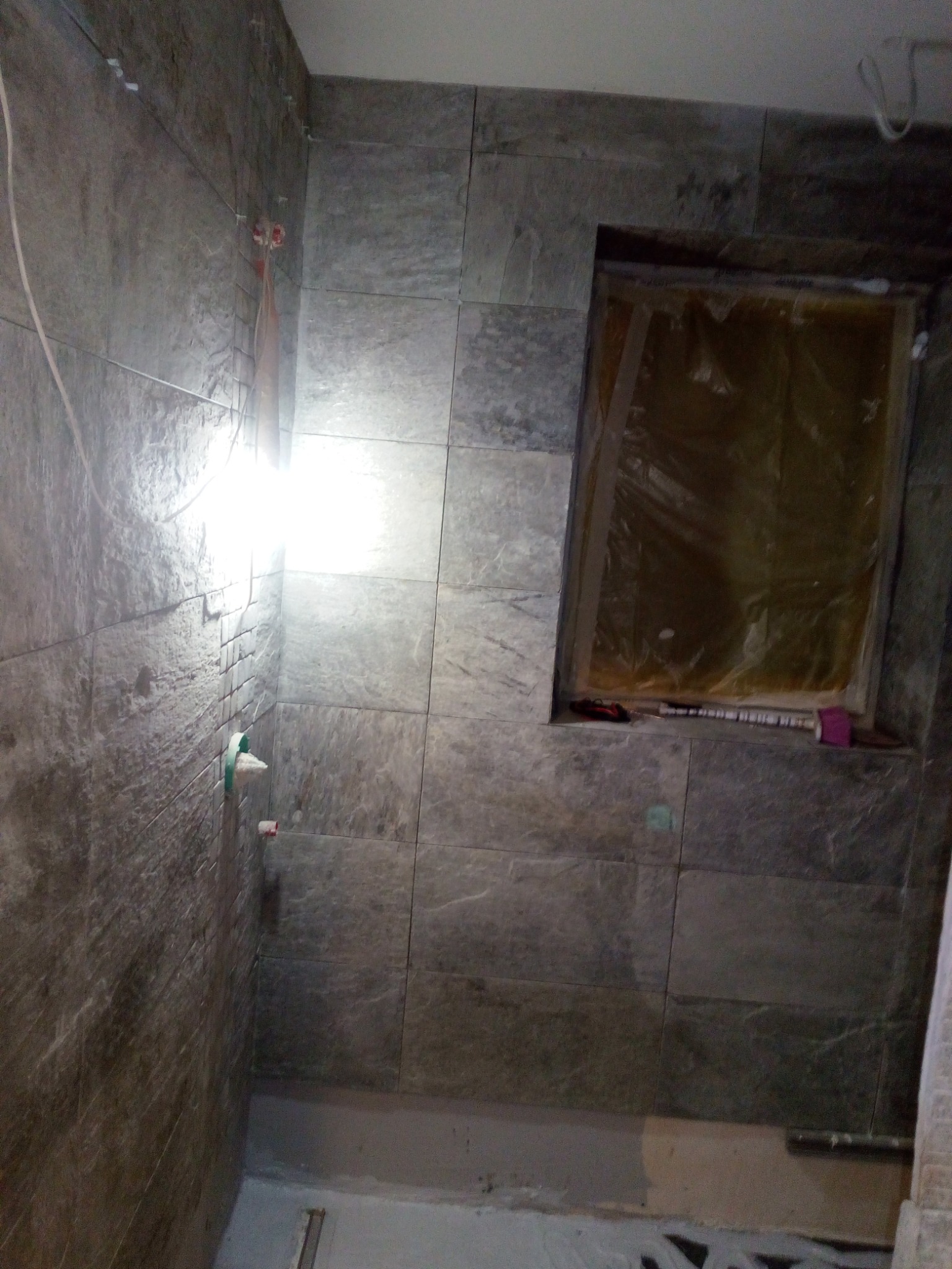 Wnętrze łazienki w trakcie remontu: szare płytki imitujące kamień na ścianach, zabezpieczone okno folią, widoczne instalacje hydrauliczne i fragment podłogi przed wykończeniem.