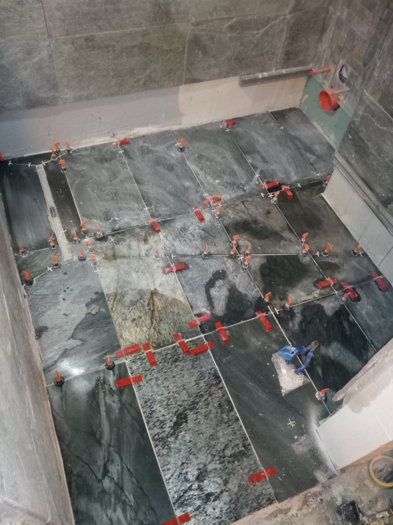 Układanie ciemnych, marmurkowych płytek podłogowych w łazience, z systemem poziomowania płytek (czerwone kliny i pomarańczowe dystanse) w trakcie prac.