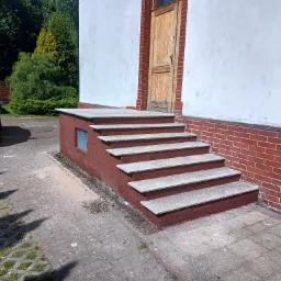 Zewnętrzne schody z szarego kamienia prowadzące do drewnianych drzwi, z boku ściana z czerwonej cegły i srebrny samochód w tle.