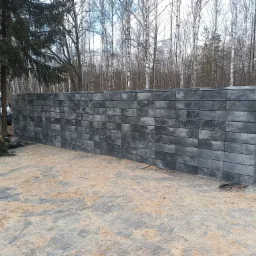 Nowo wybudowany, szary mur oporowy z betonowych bloczków, widoczny na tle drzew i częściowo zaparkowanego samochodu. Podłoże wyłożone kostką brukową pokryte piaskiem.